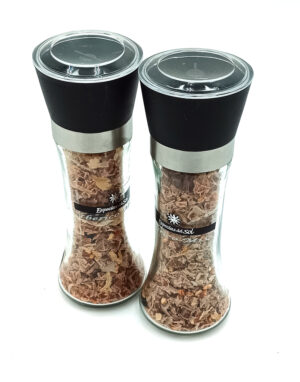 Pair of glass spice grinders with black tops and metal bands, filled with crushed brown spices (labels read 'Especiales del Sol'). They are standing side by side for seasoning food.