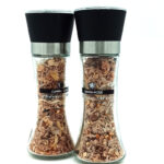 Pair of clear glass spice grinders with black tops and metal bands, filled with pinkish-brown flakes and labeled Iberico Mix.