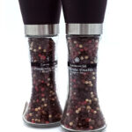 Pair of clear glass pepper mills with black tops, filled with multicolored peppercorns on a white background.