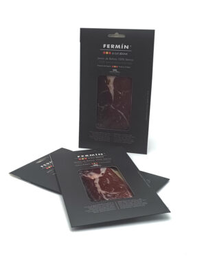 Dark packaging for Fermín 100% Ibérico ham with a transparent window showing sliced meat; several packages standing and one upright, product branding visible.