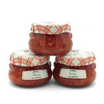 Three glass jars of tuna labeled 'Atún con fritada' with red plaid cloth lids, stacked on a white background.