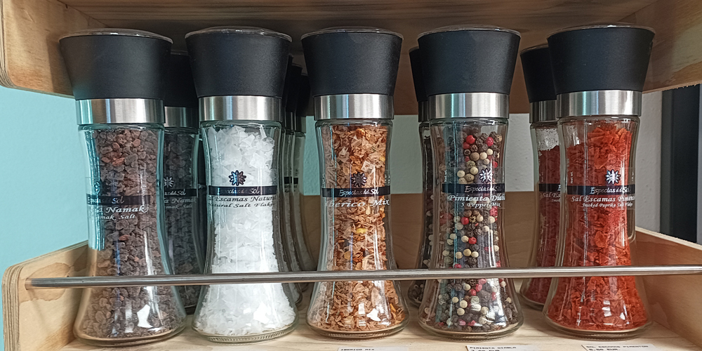 1763925976009 - El Gourmext de Casa Chino Six glass spice grinders with black tops on a wooden shelf, containing peppercorns, salt, chili flakes, and mixed spices.