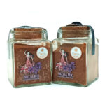 Two square glass jars with cork lids containing paella mix, labeled 'Paella Mix' with saffron and azafrán, placed side by side on a white background.