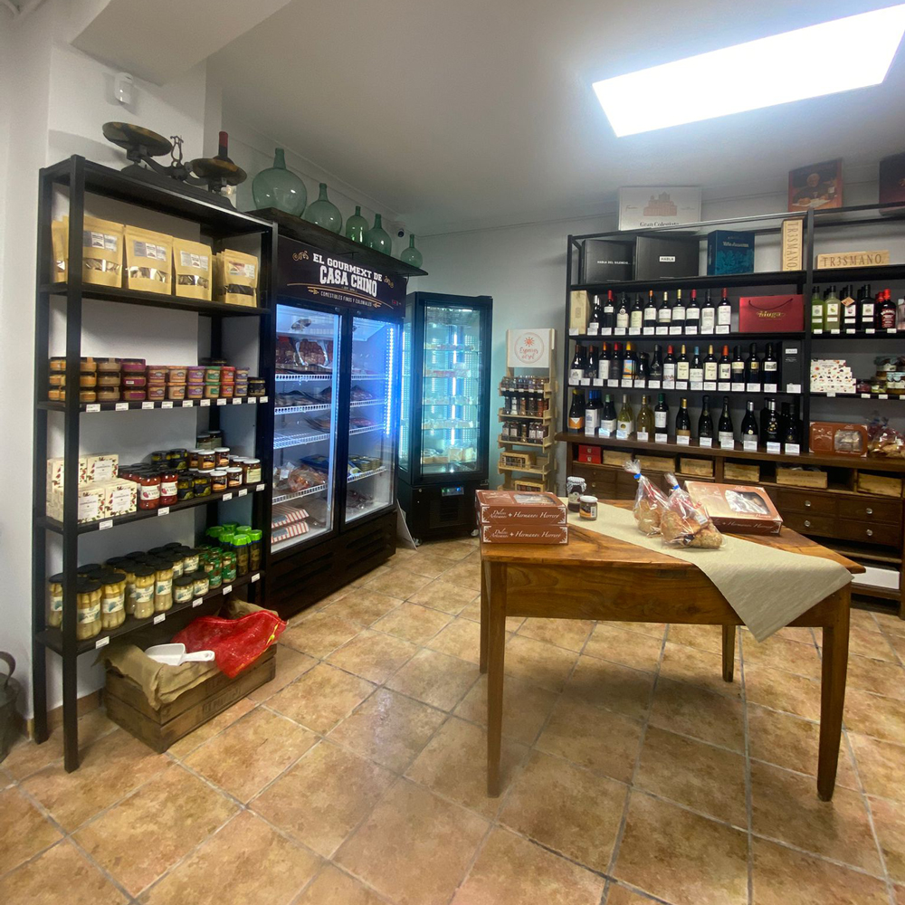 WhatsApp-Image-2025-11-01-at-101814y - El Gourmext de Casa Chino Interior of a gourmet shop with jars on shelves, wine bottles on racks, and a wooden table with baked goods in the center.
